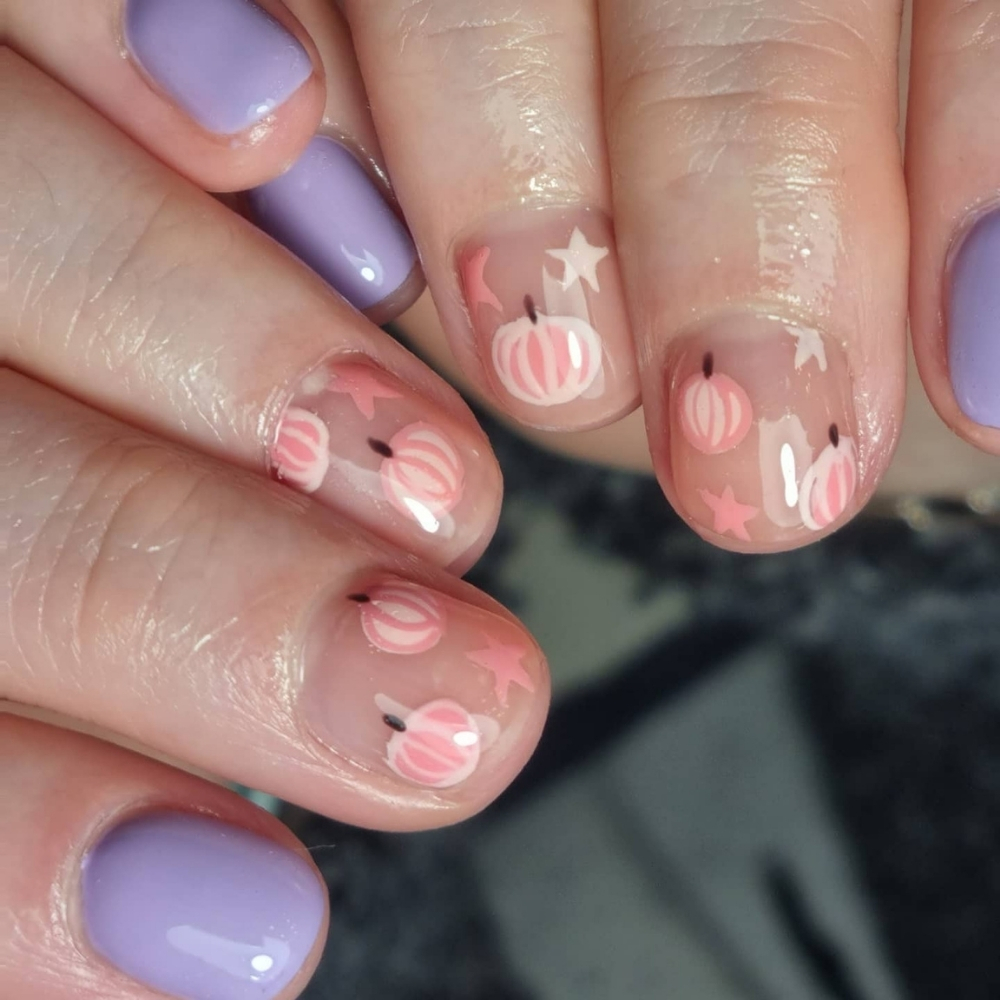 Short natural nails with clear bases, small pink and white hand painted pumpkins, tiny stars, and two lilac accent nails.