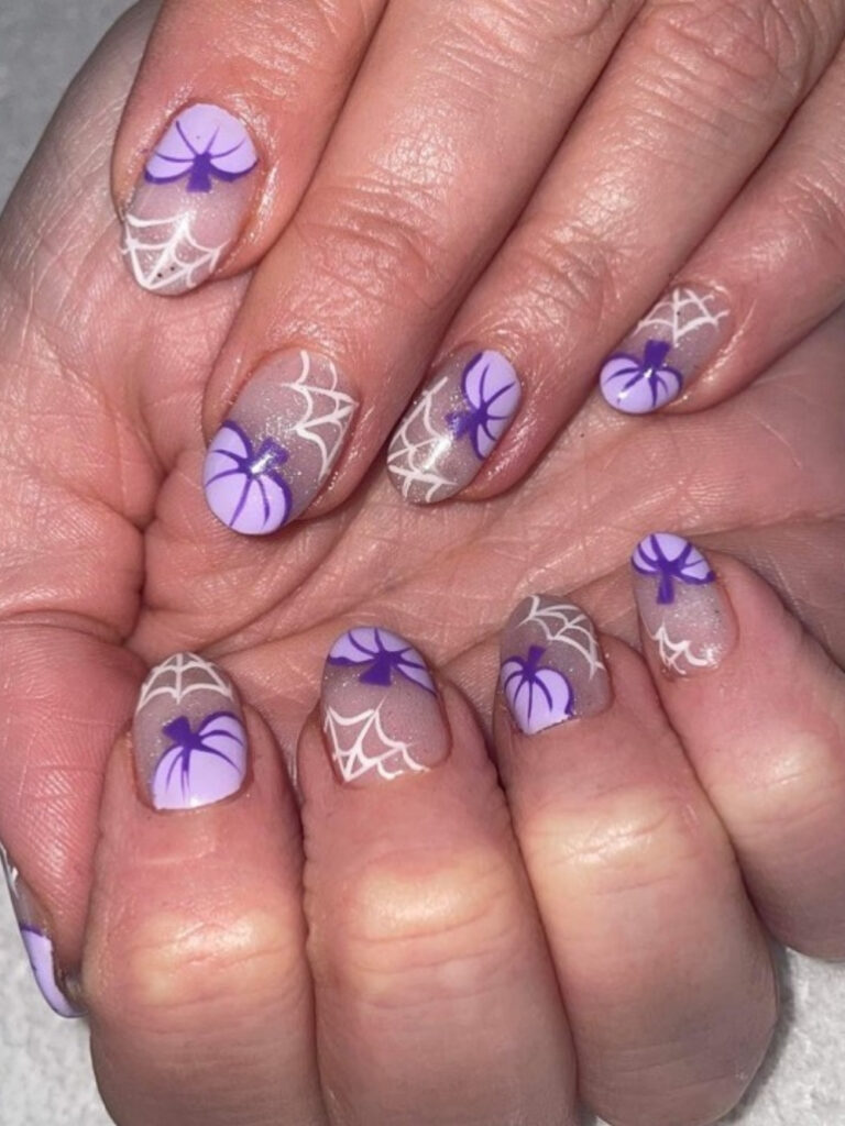 Short round nails with shimmery grey and lilac bases, hand painted purple pumpkins, and delicate white spiderwebs.