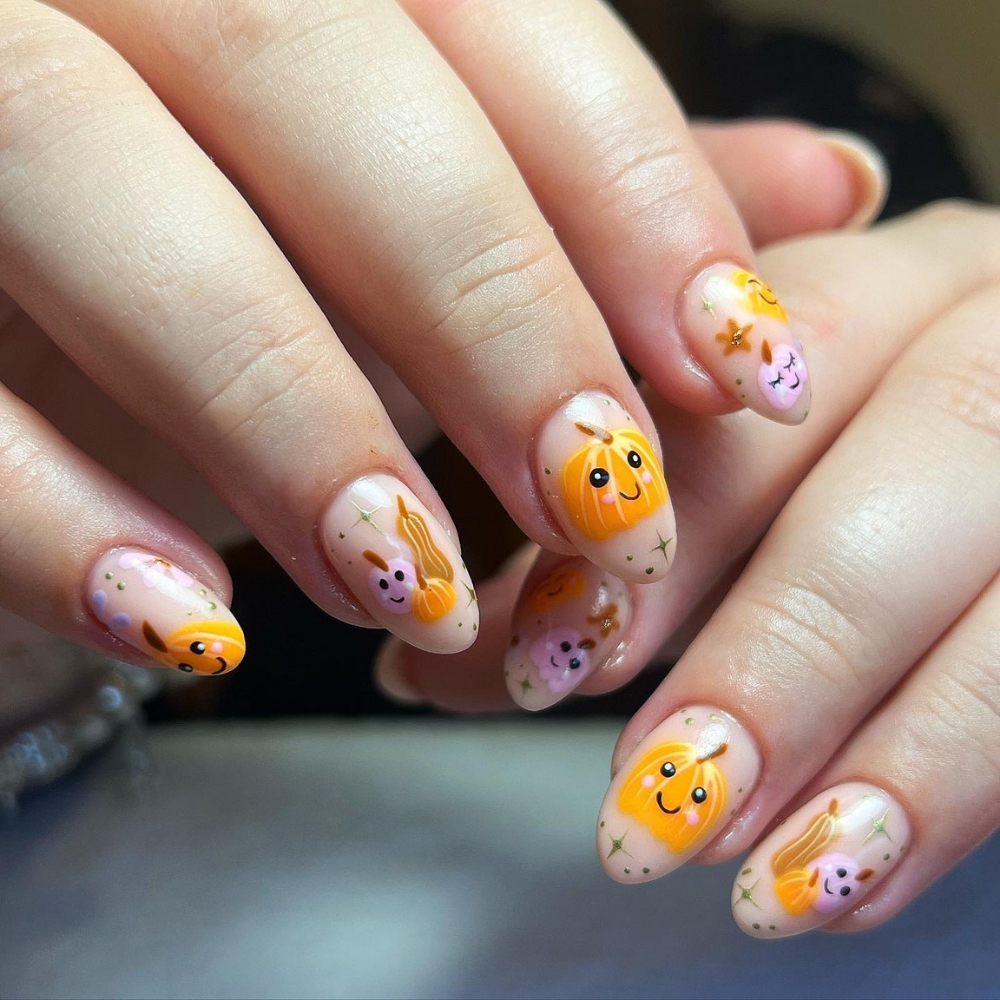 Short almond nails with sheer nude bases featuring hand painted smiling orange and pink pumpkins with gold sparkles and dots.