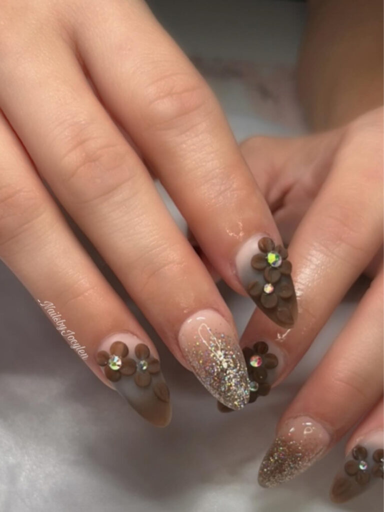 Long almond nails with sheer nude bases, raised brown 3D flowers with crystal centers, and champagne glitter ombre accents.