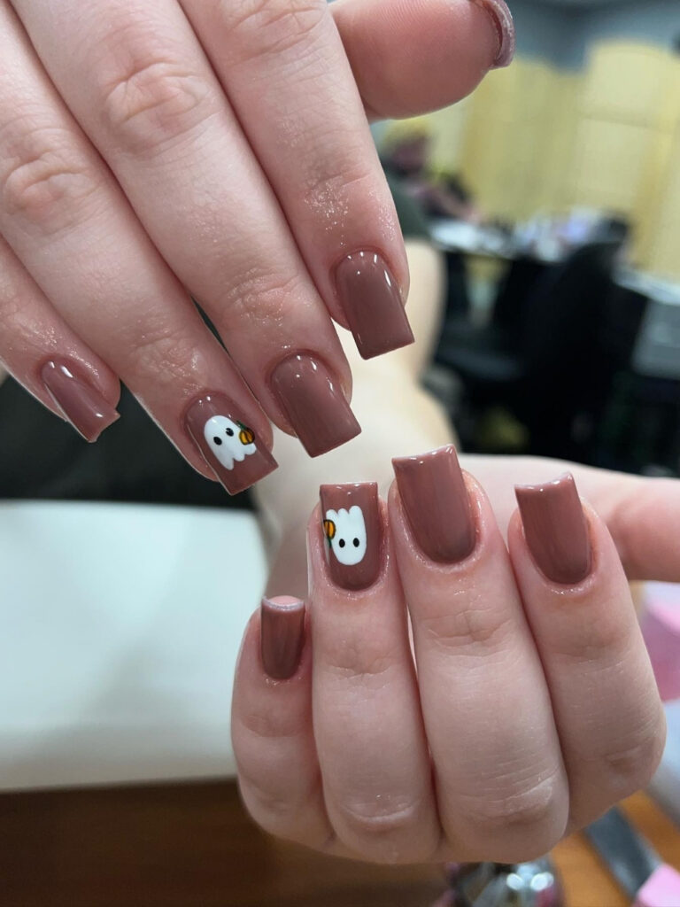 Cute fall nails on medium square tips in warm mocha brown, with a single accent nail featuring a small white ghost holding a tiny pumpkin.