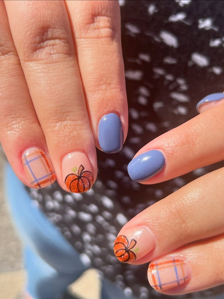 Short round nails in periwinkle blue with sheer accent nails featuring orange plaid and a glitter pumpkin.