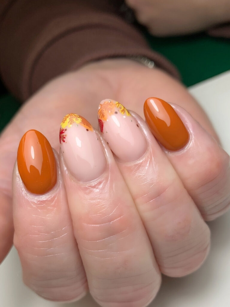 Medium almond nails in glossy burnt orange with sheer nude accent nails featuring stamped autumn leaves in gold, red, and orange.