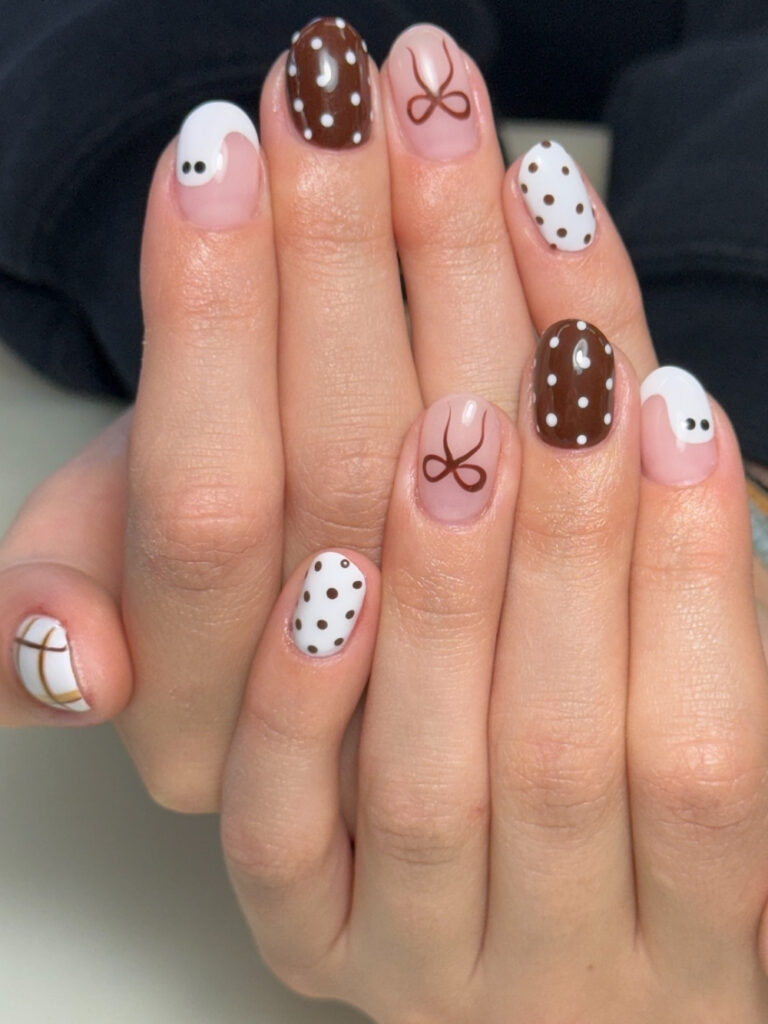 Short squoval nails mixing brown polka dots on white, white dots on brown, white swoosh nails with dot detail, hand painted brown bows, and a plaid accent.