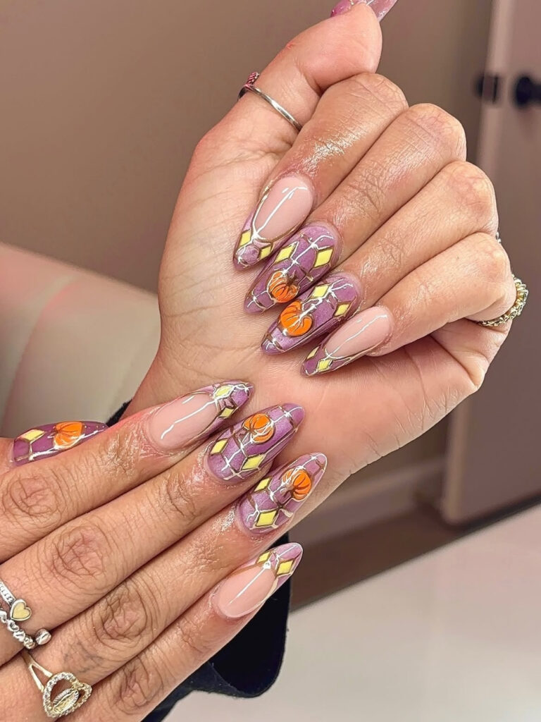 Long almond nails in shimmery lilac with gold outlined diamond patterns, and hand painted orange pumpkins.