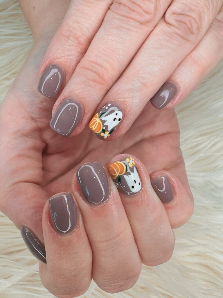 Cute brown fall nails on short square tips in muted taupe, with a single accent nail featuring a white ghost, a small orange pumpkin, and yellow flowers.