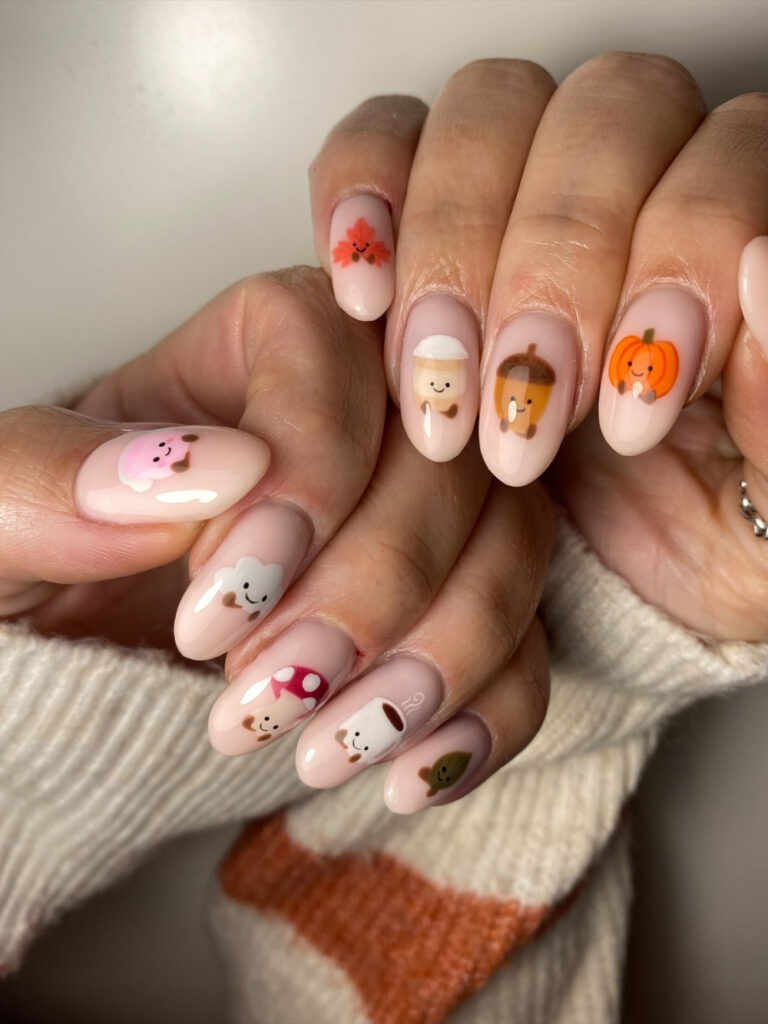 Autumn nails on medium almond tips with sheer pink bases, featuring smiling hand painted pumpkins, acorns, mushrooms, maple leaves, and a latte cup.
