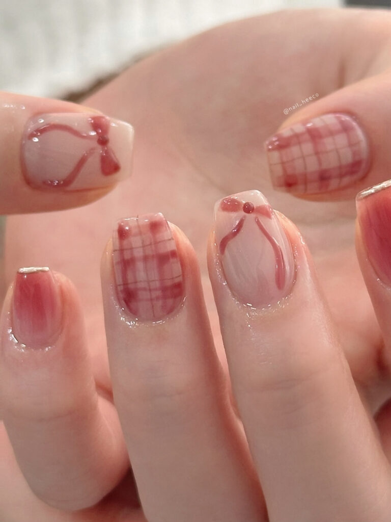 Short square nails with milky bases, dusty rose plaid accents, and delicate raised ribbon bows in a matching red tone.
