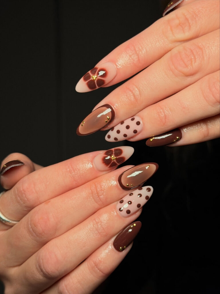 Long almond nails in milky white and rich brown with oversized polka dots, blurred floral accents, and gold micro studs.