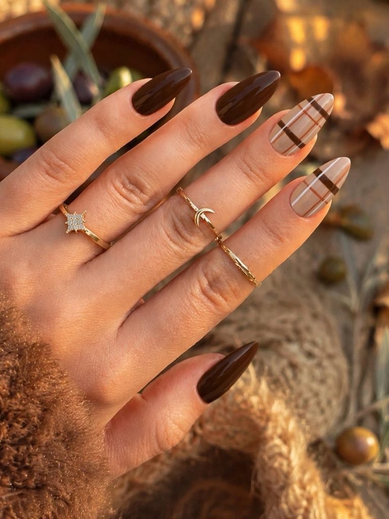 Long almond nails in glossy dark chocolate brown with two accent nails in sheer nude plaid, styled with gold rings in golden hour light.