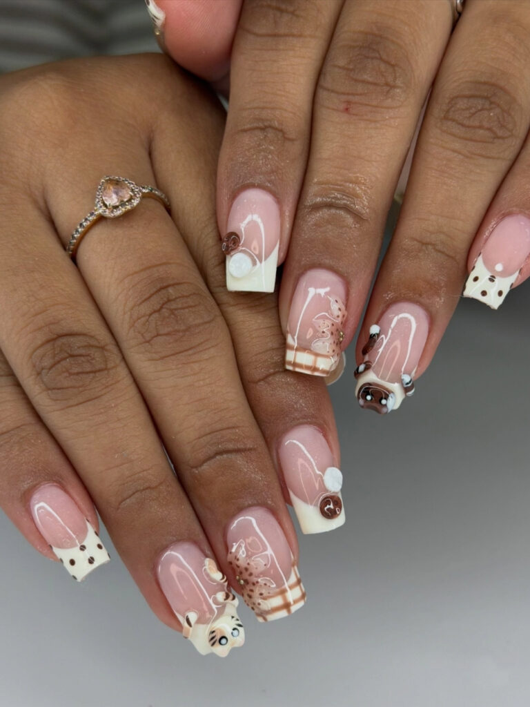Long square nails with sheer pink bases and cream French tips, featuring brown polka dots, gingham and leopard print accents, and 3D cat face charms in cocoa and striped cream. 