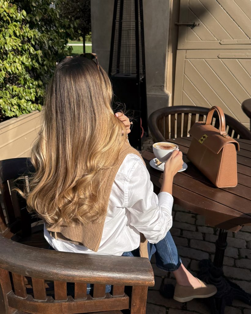 Woman with golden honey blonde balayage hair sipping coffee outdoors, a warm fall blonde hair color idea with soft, sun-kissed dimension.
