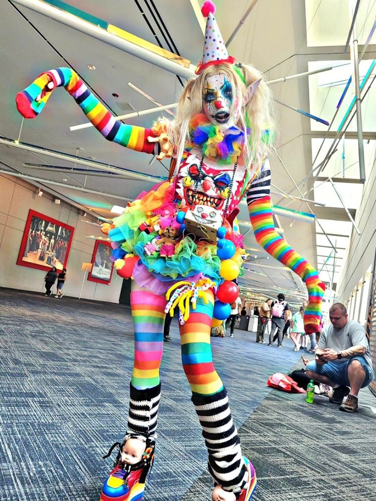 Elaborate avant-garde clown outfit with rainbow-striped sleeves and dramatic makeup.