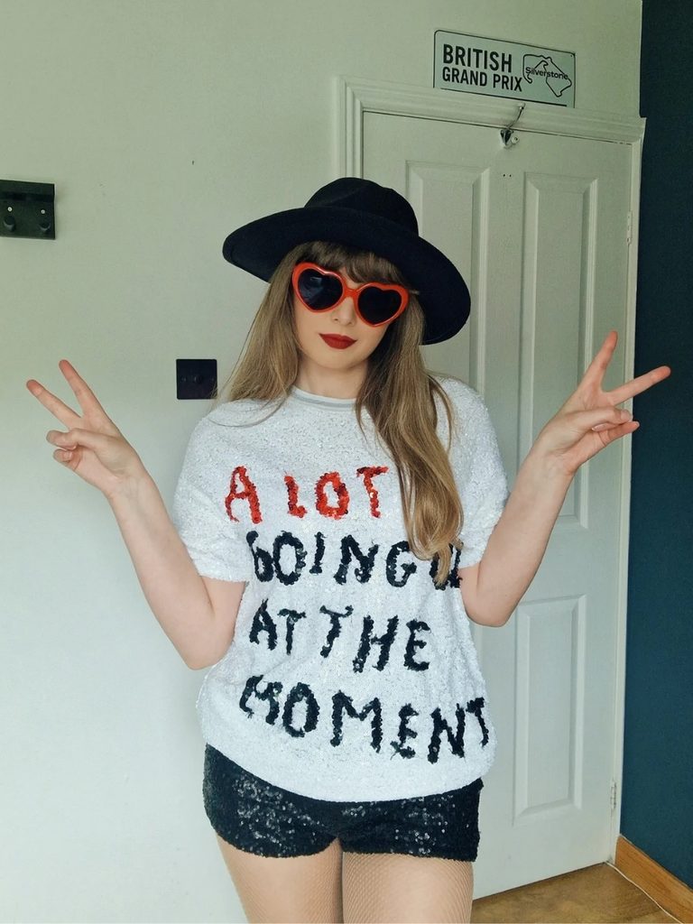 Person wearing a graphic tee, sequin shorts, heart-shaped sunglasses, and a wide-brim hat. This Halloween costume is inspired by Taylor Swift.
