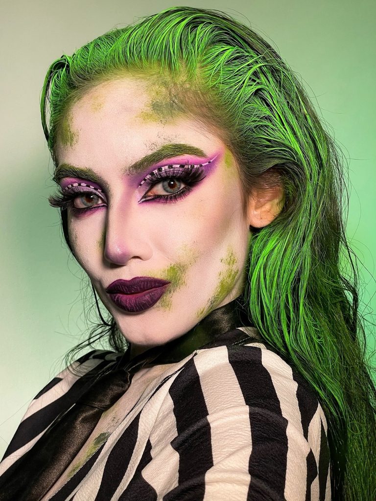 Slicked green hair, checkered lilac eyeshadow, and dark plum lipstick create this Beetlejuice-inspired Halloween makeup look.