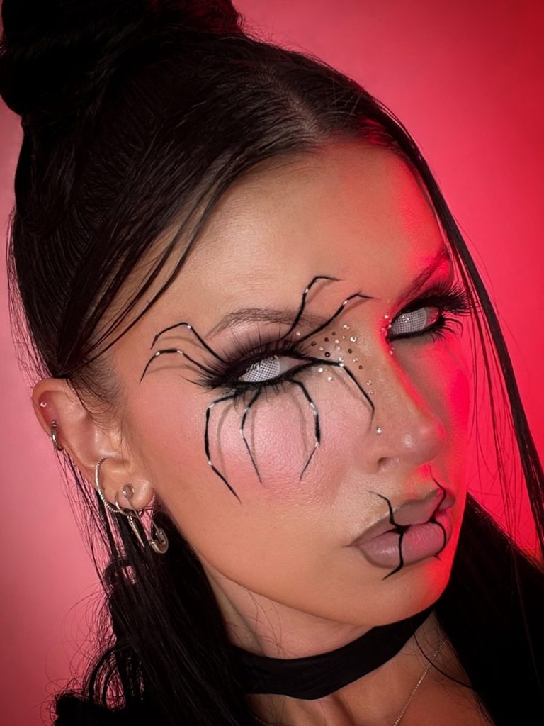 Long black spider legs drawn around the eye and scattered rhinestones create dramatic Halloween eye makeup.