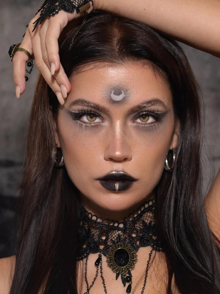 A hand-painted crescent moon on the forehead, smoky eyes, and black lipstick create a mystical witch makeup look.