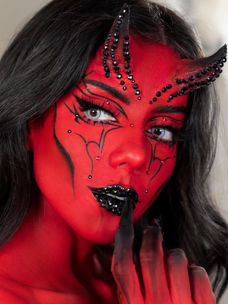 Full red face paint, sculpted devil horns, and black crystals along the lips create a bold and creative Halloween makeup look.