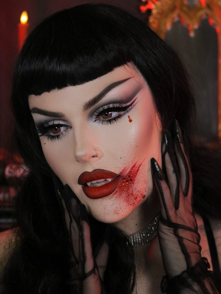 A metallic smoky eye, sharp fangs, and a blood detail at the mouth create a dramatic vampire makeup look.