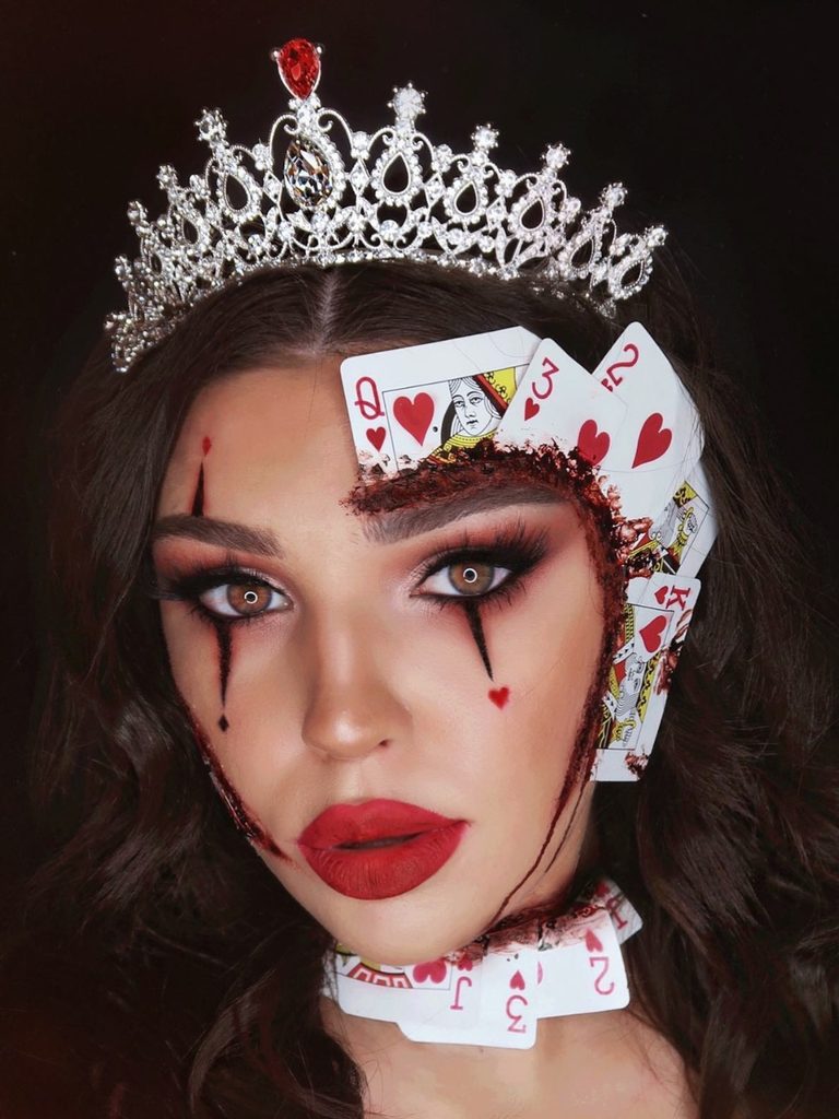 Playing cards arranged around a rhinestone tiara, heart-shaped eyeliner, and a dripping red brow create a creative Halloween makeup look.