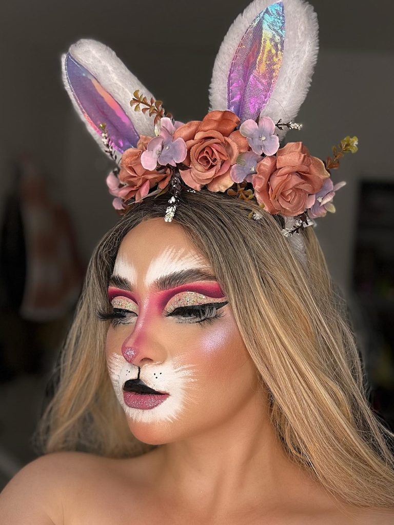 Pink and gold eyeshadow, a painted nose, white whiskers, and a floral ear headband create pretty bunny makeup.