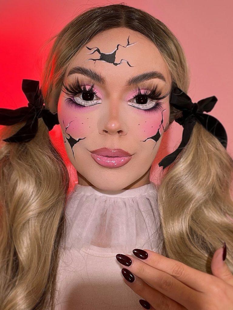 Painted fracture lines across the forehead and cheeks, oversized lashes, and rosy blush create a creepy broken-doll effect.