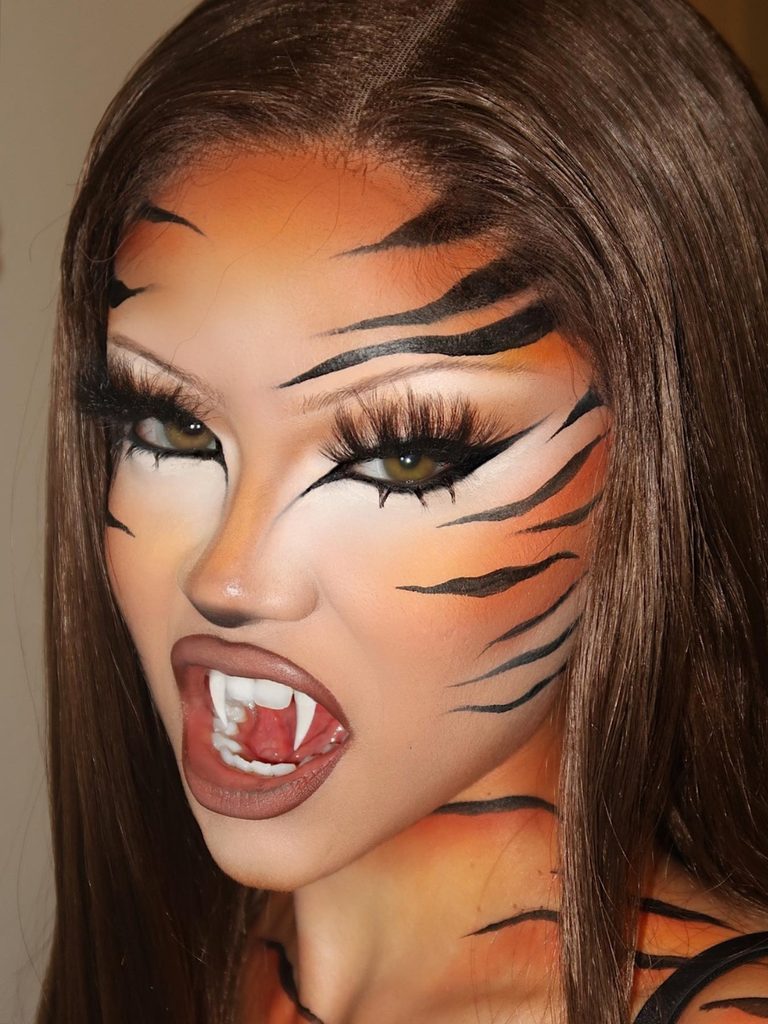 Bold black stripes over burnt-orange face paint and dramatic fangs bring this tiger-inspired Halloween face makeup to life.