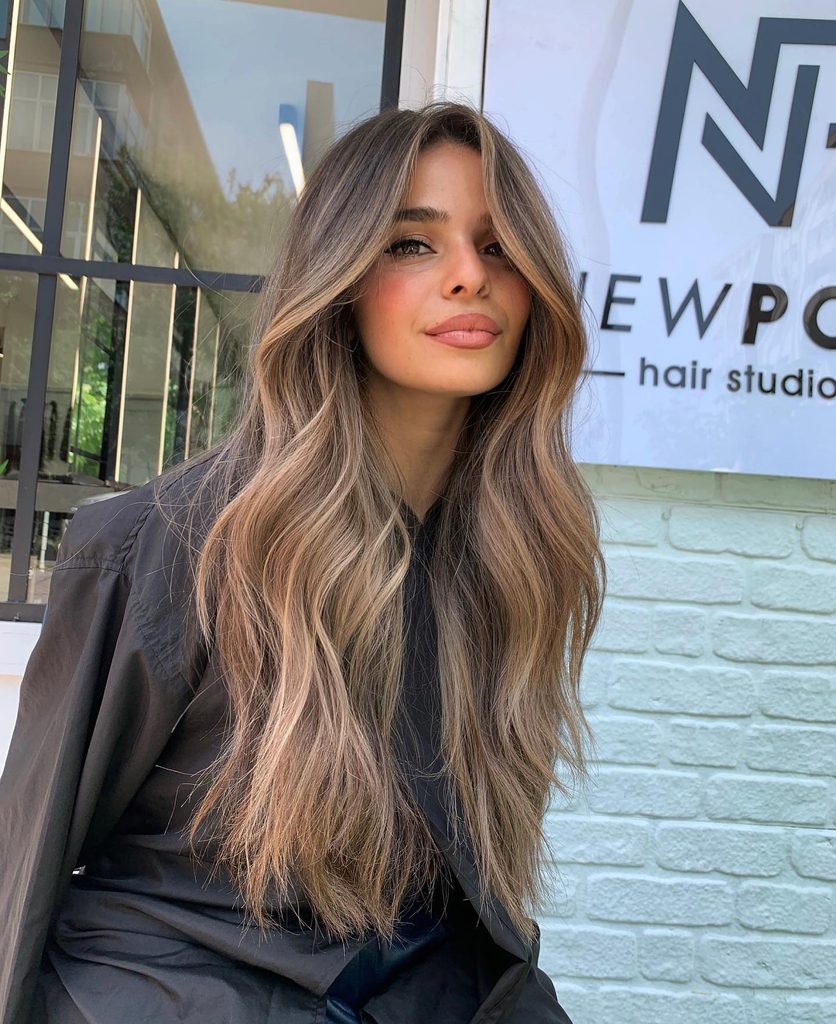 Woman with long wavy caramel bronde balayage hair standing outside a salon, a low-maintenance fall hair color idea for blondes.