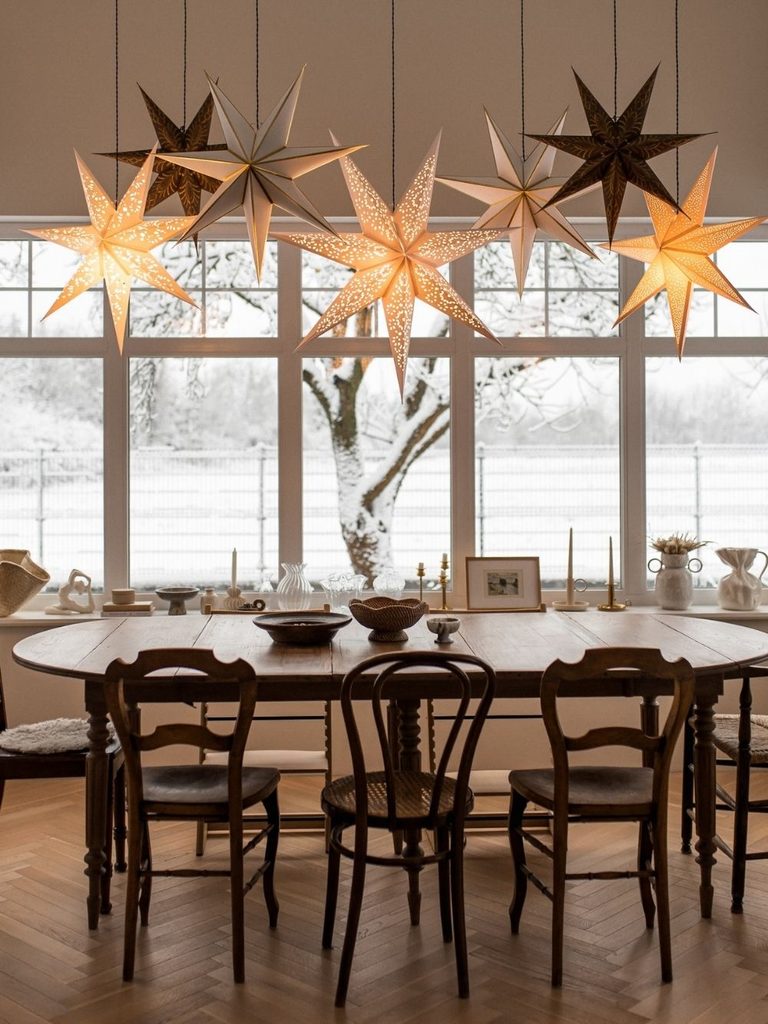 Neutral dining room with paper star lanterns hanging above a wood table in front of snowy windows