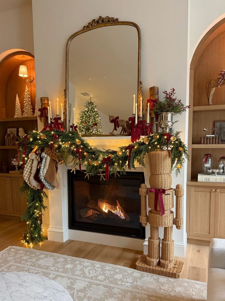 Christmas mantel decor with garland, stockings, and a rattan nutcracker beneath a large gold mirror