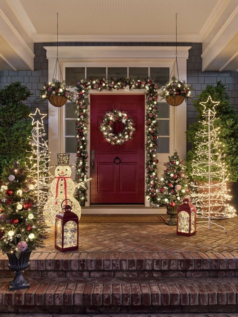 Front porch Christmas decor with lit spiral trees, a snowman, lanterns, and a wreath on a red door
