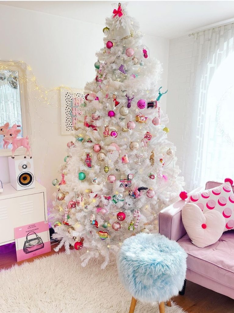 White Christmas tree with pastel and rainbow retro ornaments beside a pink sofa and fuzzy stool