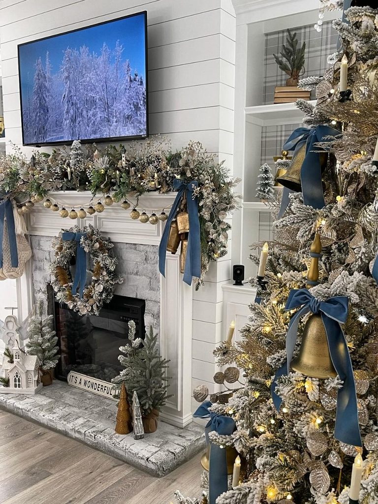 Christmas mantel decor with frosted garland, blue velvet bows, gold bells, and a wreath at the hearth beside a flocked tree