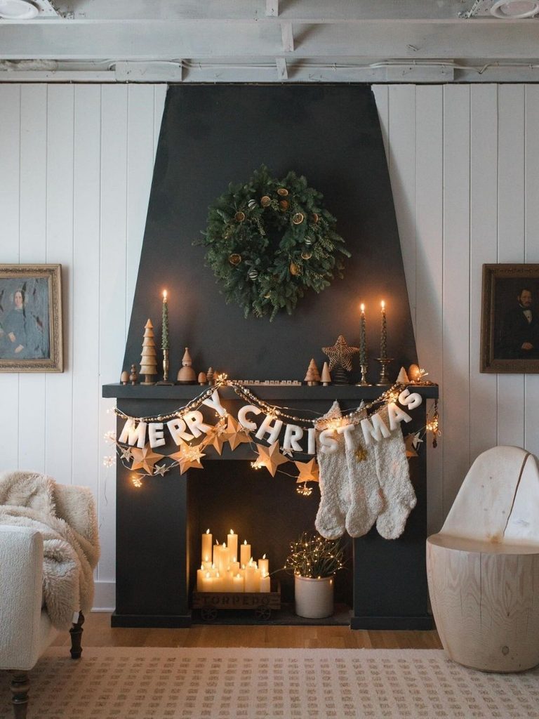 Black fireplace with a wreath, a letter Merry Christmas banner, stockings, and candles glowing in the hearth