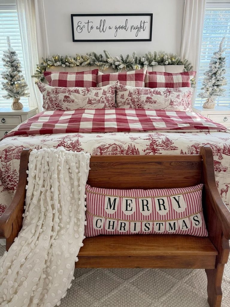 Bed with red and white gingham and toile bedding, a Merry Christmas pillow on a wooden bench, and mini trees