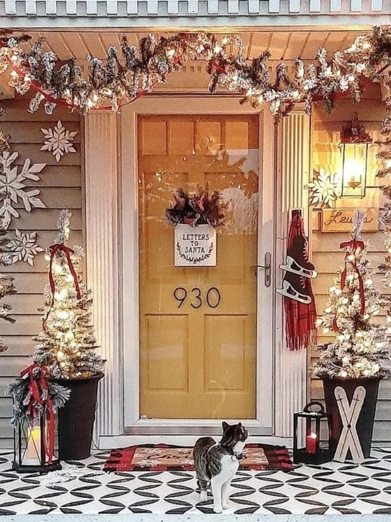 Porch with a yellow door, flocked trees in planters, lanterns, light-up garland, and a cat on a patterned tile floor