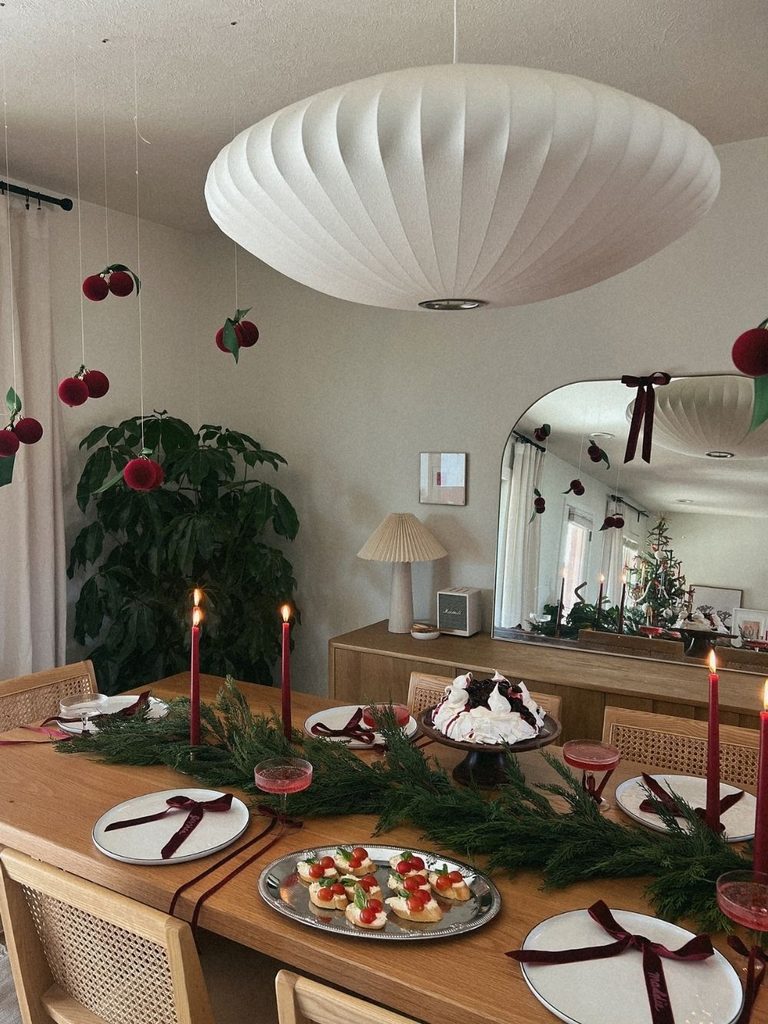 Dining table with an evergreen garland runner, red taper candles, velvet bow place settings, and burgundy velvet cherries hanging from the ceiling