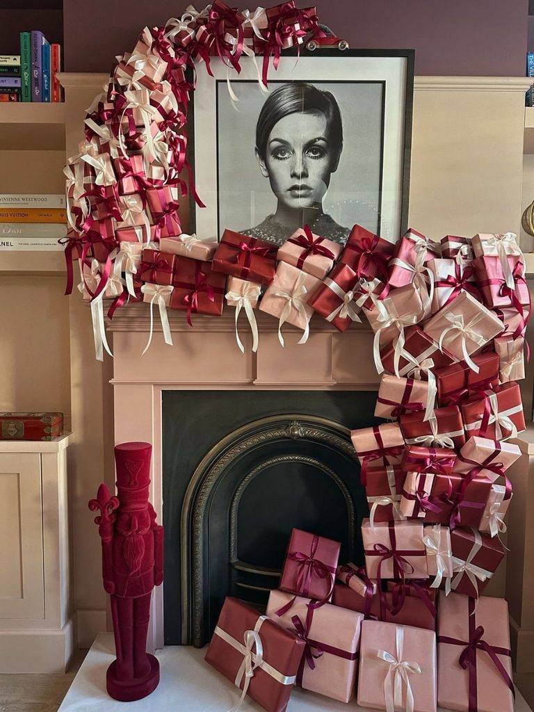 Pink fireplace mantel stacked with red, blush, and burgundy wrapped presents beside a nutcracker