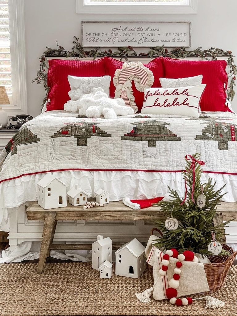 White bedroom with a Christmas tree quilt, red and white pillows, a wooden bench, and a mini tree and white houses at the foot of the bed