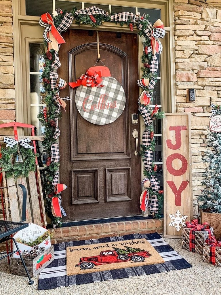 Wood front door with plaid garland, a buffalo check ornament, a JOY sign, a sled, and a red truck doormat