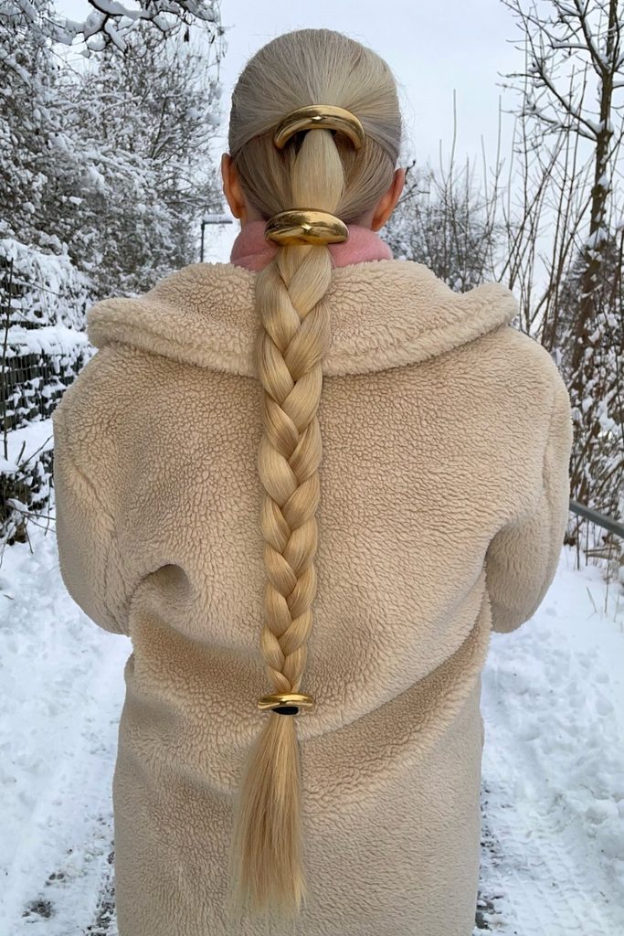 Sleek winter hair braid with gold cuffs worn over a cream shearling coat in the snow